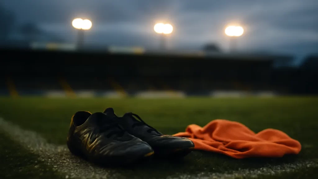 Scuffed football boots and plain bib on wet pitch under floodlights.